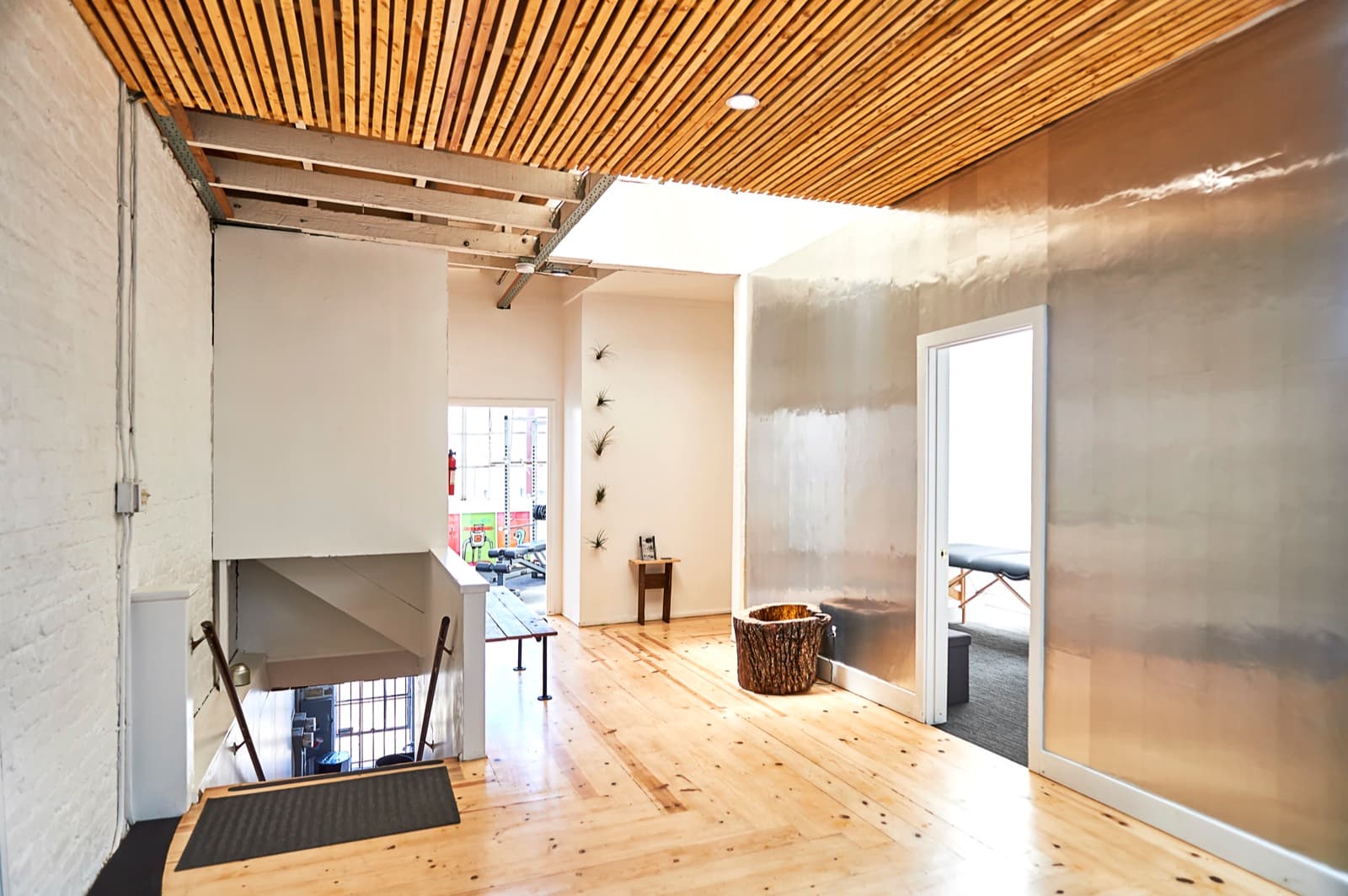 Wellness center after the fit-out from the same spot: oak floor, white brick, wood-slat ceiling, a mirror wall and suspension straps