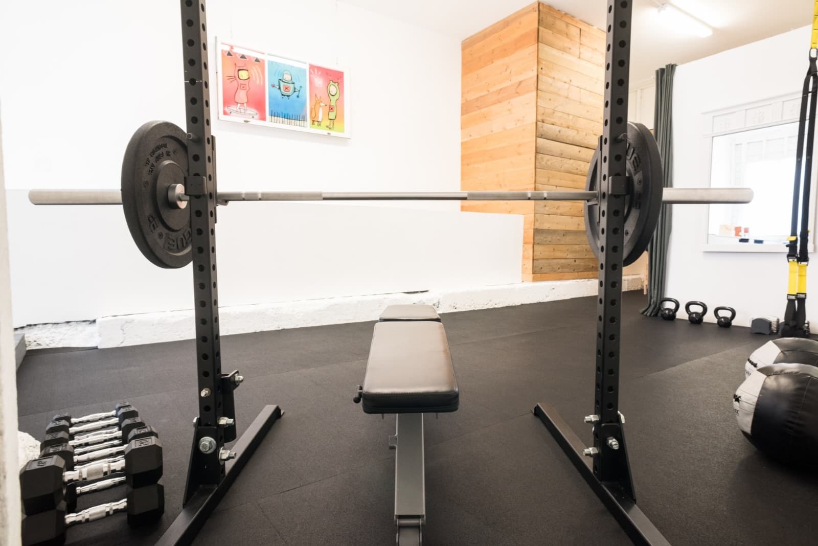 Guerrero Street studio after: a squat rack with a loaded barbell, a flat bench and dumbbells on a black rubber floor, a cedar column and a window behind