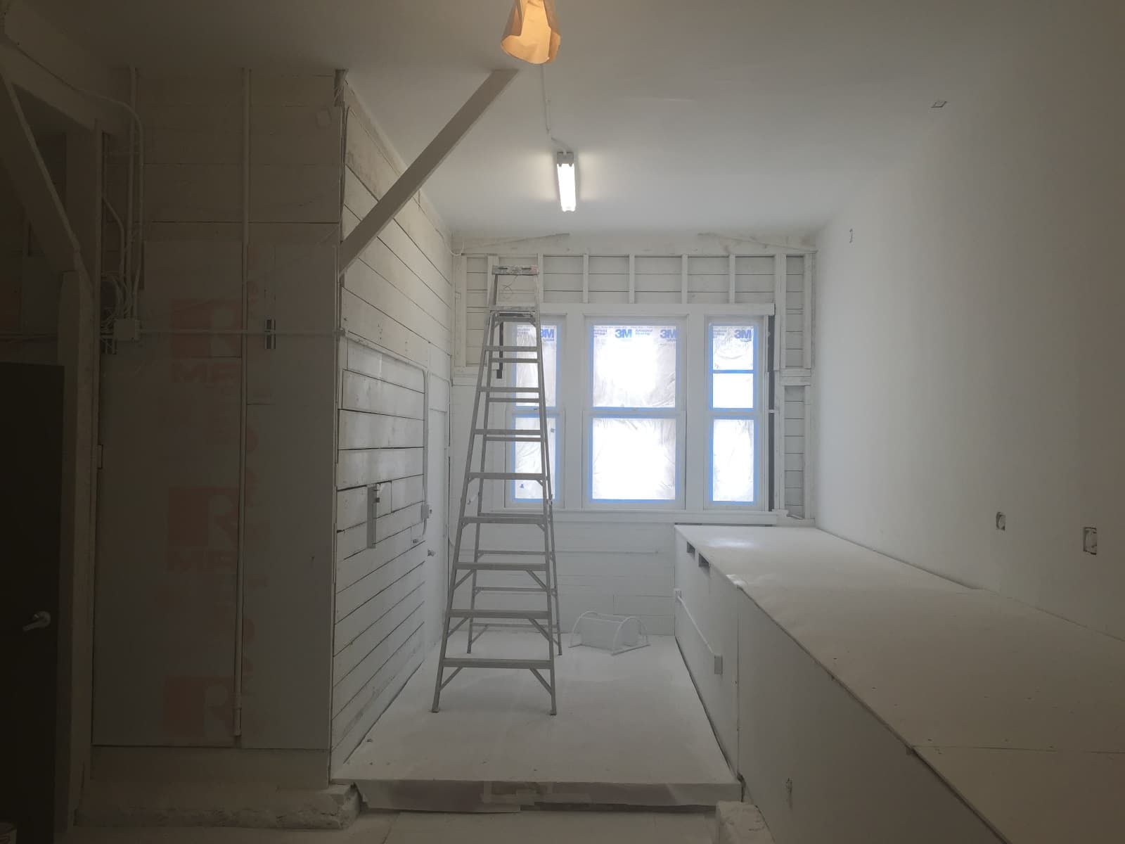 Guerrero Street garage mid-conversion, looking toward the street window: first coat of white on the walls and ceiling, a stepladder, a built-in bench frame at right and a fluorescent light overhead