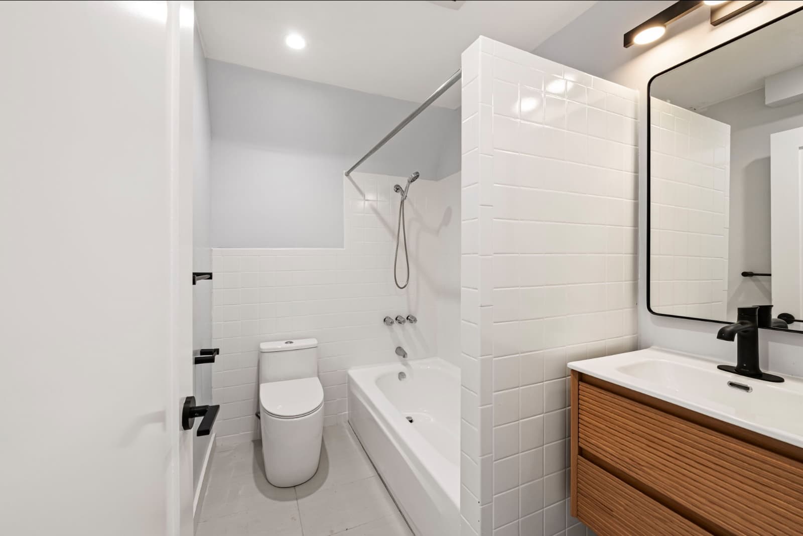 Corona Heights home downstairs bathroom after the remodel: white tiled tub with a glass panel, a wood vanity, black fixtures and a black-framed mirror